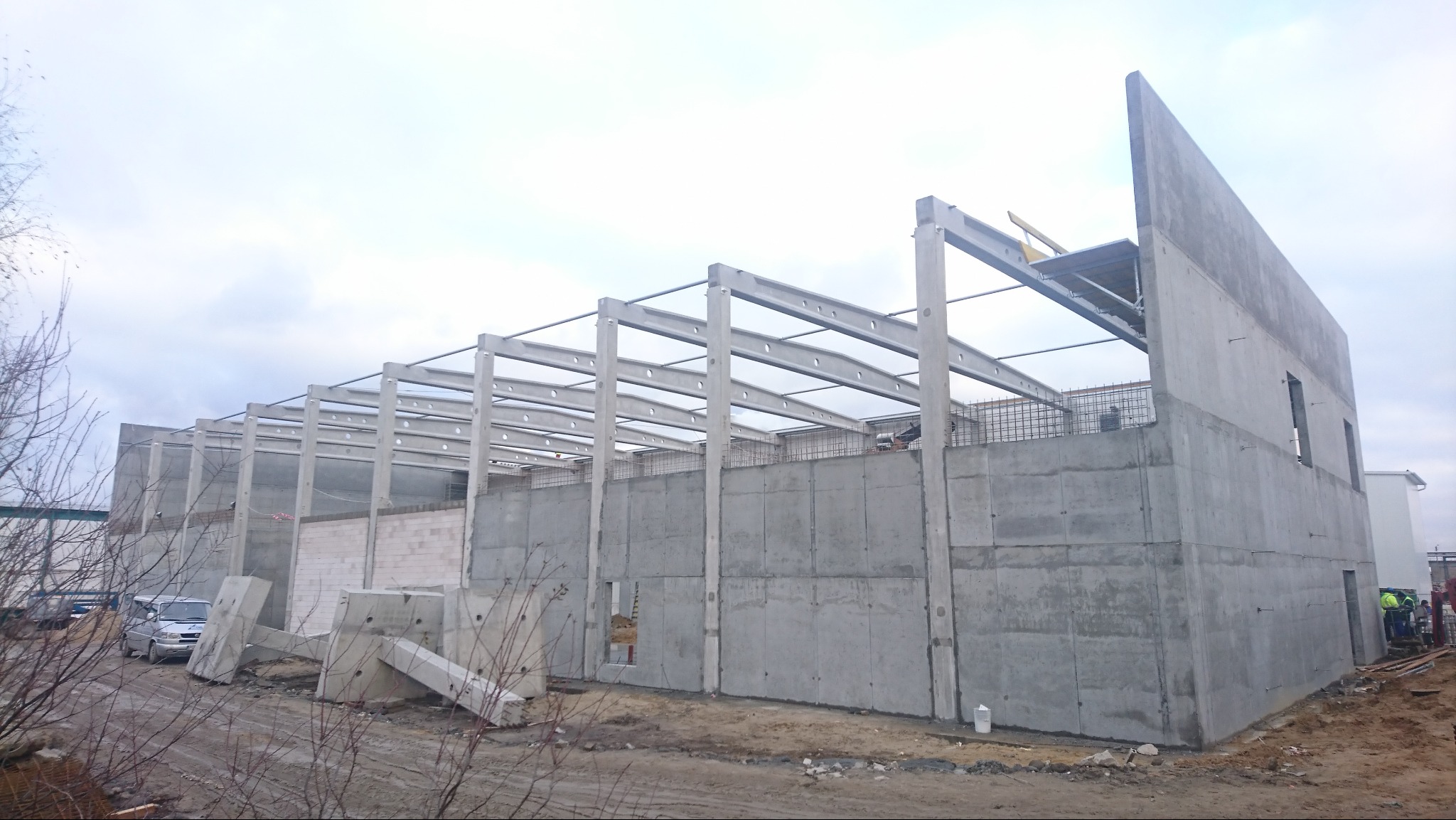Budowa hali przemysłowej z prefabrykatów betonowych, widoczne słupy i ściany w surowym stanie, elementy konstrukcji stalowej na dachu, na ziemi leżą betonowe bloki, w tle zaparkowany samochód...