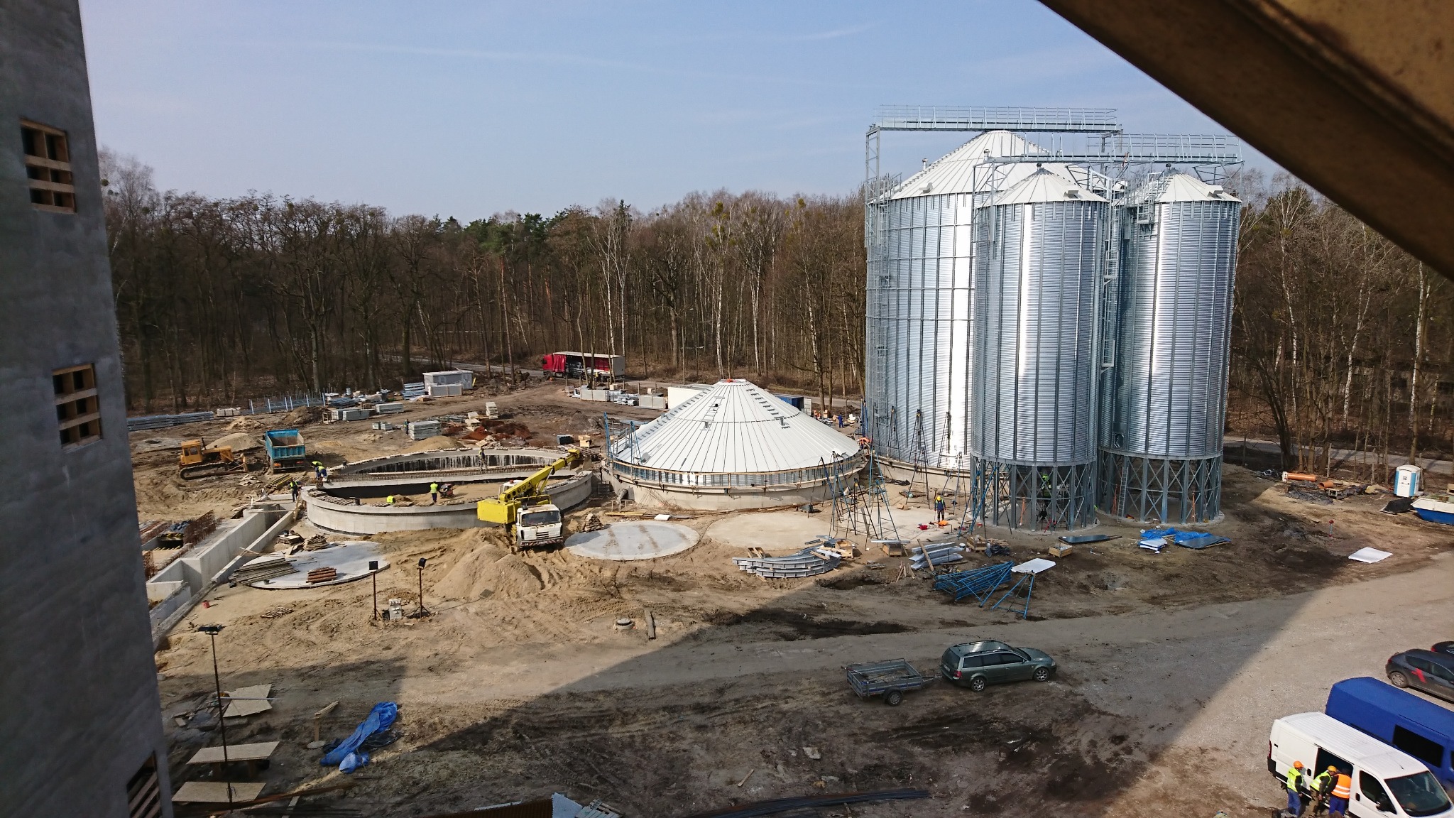 Budowa oczyszczalni ścieków z widokiem na okrągły zbiornik z kopułą i wysokie, metalowe silosy, w tle las i plac budowy z maszynami.