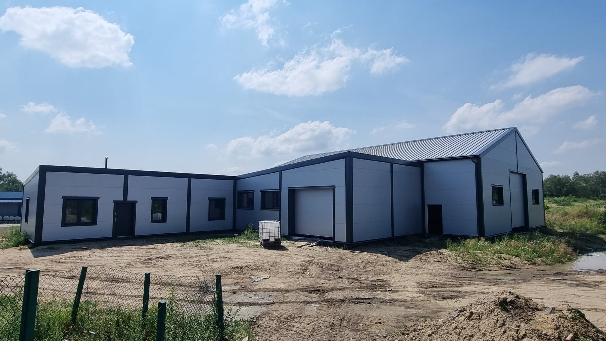 Nowoczesna hala magazynowa z szarej blachy trapezowej, widok z zewnątrz, jasne niebo z chmurami w tle, teren wokół niezagospodarowany.