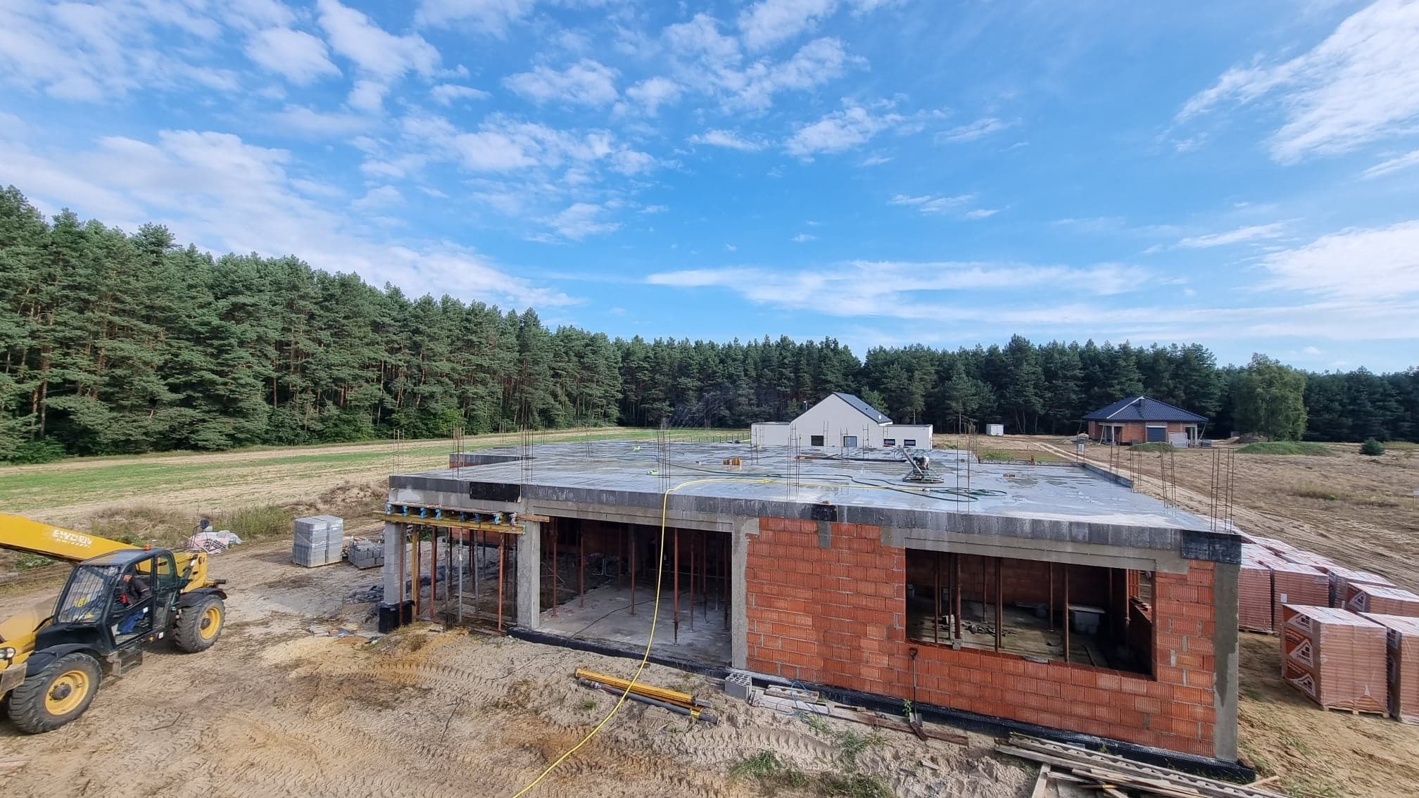 Budowa domu jednorodzinnego w stanie surowym otwartym, widoczne ściany z czerwonej cegły, betonowy strop z wystającymi prętami zbrojeniowymi, podpierające konstrukcje, na pierwszym planie ładowarka...