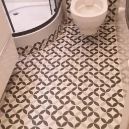 Wykończona łazienka z kabiną prysznicową i toaletą, wzorzyste płytki podłogowe i ścienne w odcieniach szarości, widok z góry, częściowo widoczny parkiet podłogowy.