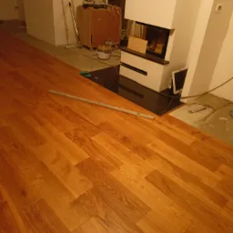 Układanie paneli podłogowych w pomieszczeniu z kominkiem, widoczna część ułożonej podłogi i narzędzia w tle.