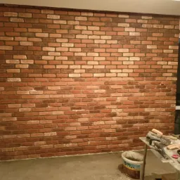 Ściana wyłożona dekoracyjną cegłą w odcieniach brązu i beżu, widoczne narzędzia i wiadro z zaprawą na stole, okno w tle.