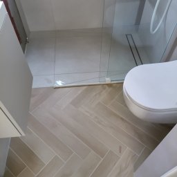 WR Remont dla Ciebie - Wykończona łazienka z beżową podłogą ułożoną w jodełkę, białą toaletą i szklaną kabiną prysznicową. Minimalistyczny design, neutralna kolorystyka.