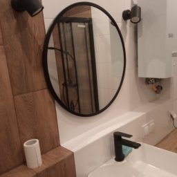 WR Remont dla Ciebie - Nowoczesna łazienka z okrągłym lustrem w czarnej ramie, białą umywalką z czarną armaturą i drewnopodobnymi płytkami. Widoczny bojler i oświetlenie.