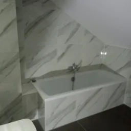 Biała wanna obudowana płytkami imitującymi marmur w łazience na poddaszu, widoczny fragment toalety i ciemna podłoga.