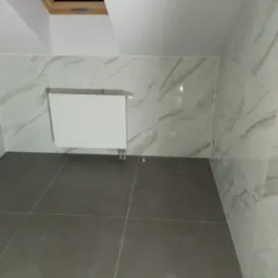 Wnętrze łazienki na poddaszu z szarymi płytkami podłogowymi i marmurkowymi płytkami ściennymi, biały grzejnik drabinkowy i okno dachowe.