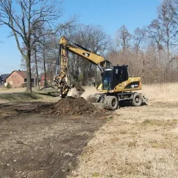 Żółta koparka CAT nabiera ziemię z wykopu na tle wiejskich domów i drzew bez liści, obok stos drewna.