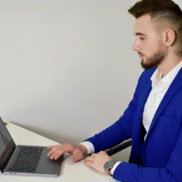 Mężczyzna w niebieskiej marynarce siedzi przy biurku i pracuje na laptopie, na ekranie widoczna strona internetowa z wizerunkiem osoby.