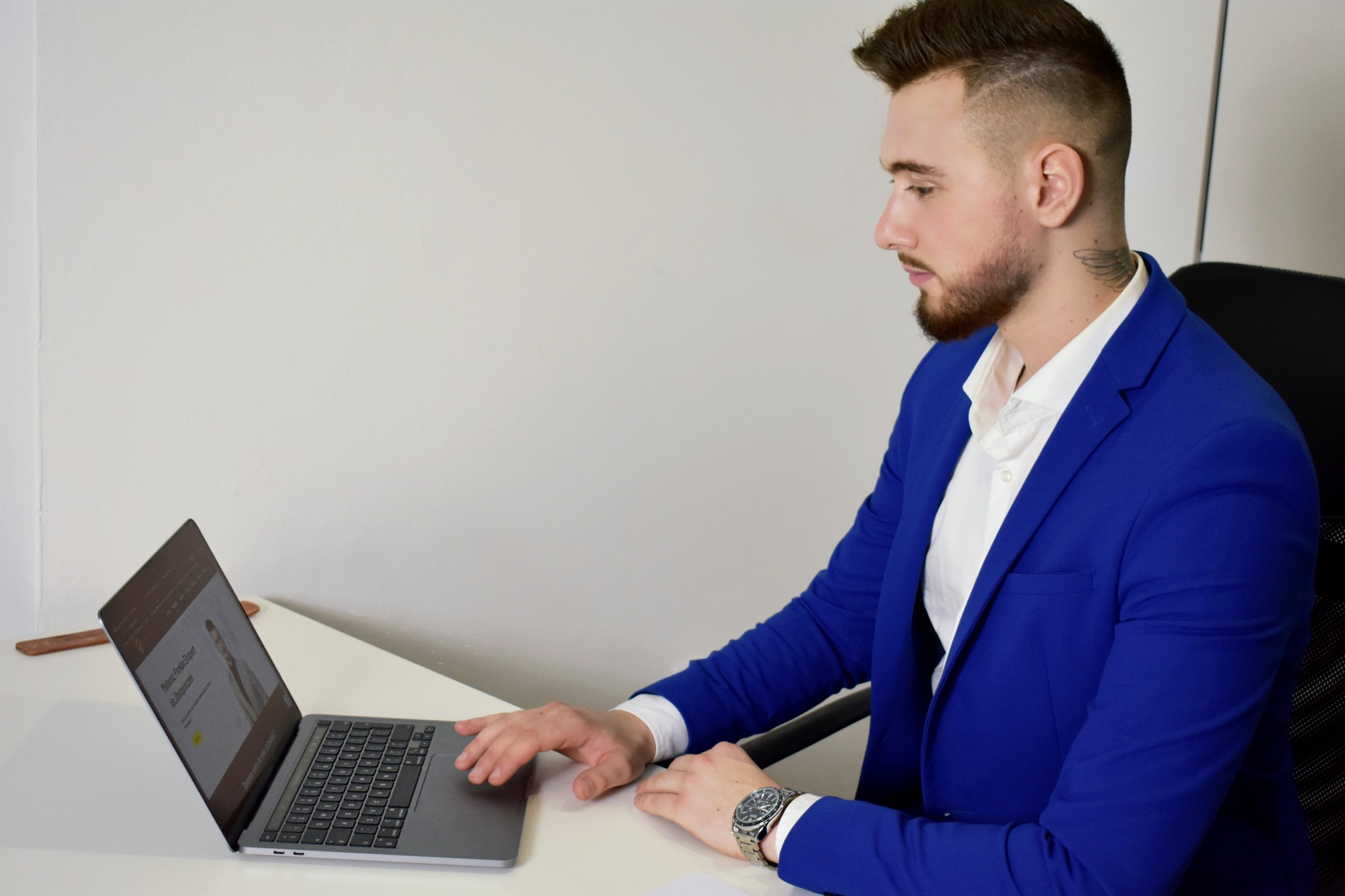 Mężczyzna w niebieskiej marynarce siedzi przy biurku i pracuje na laptopie, na ekranie widoczna strona internetowa z wizerunkiem osoby.