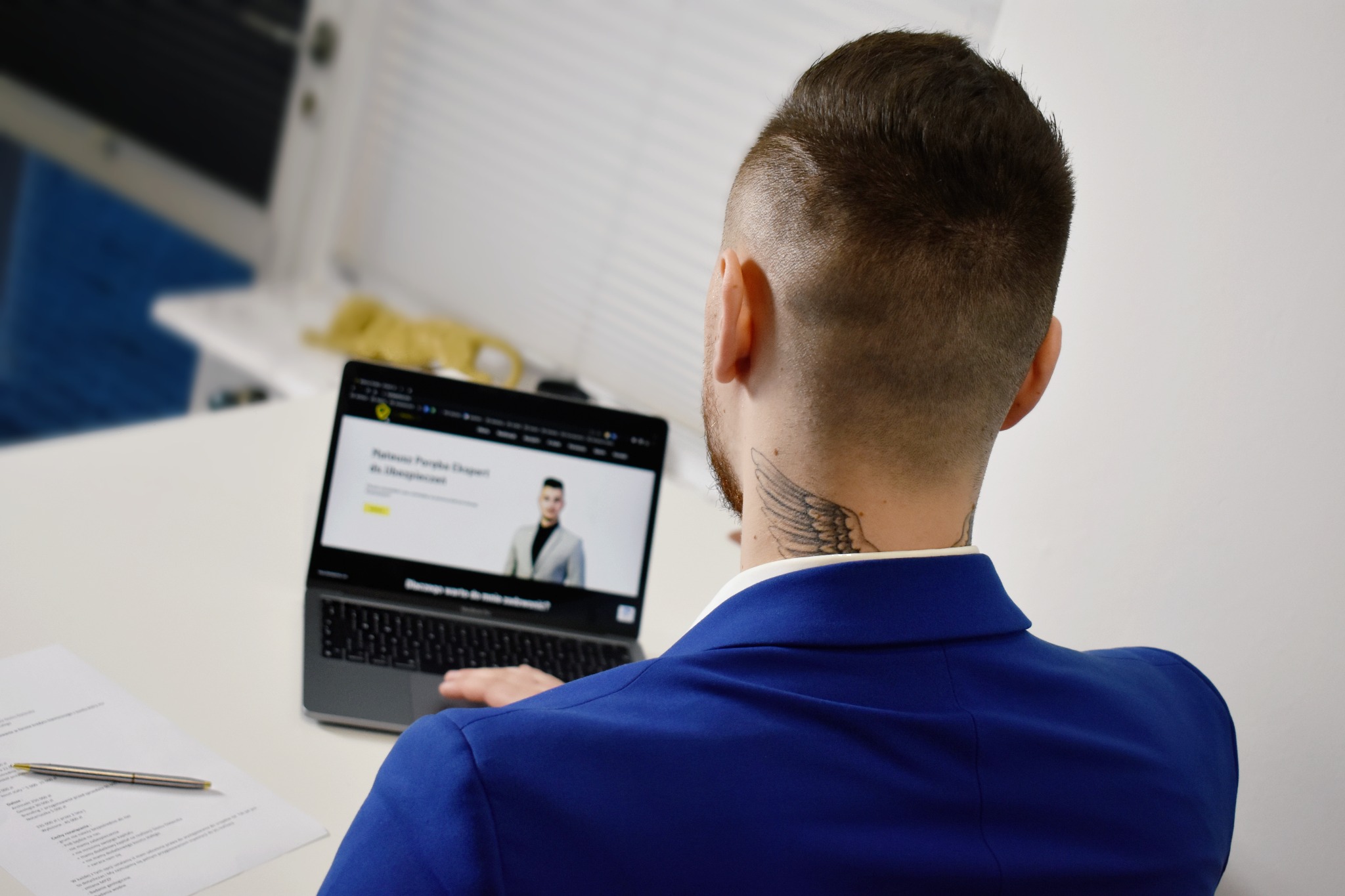 Mężczyzna w niebieskiej marynarce z tatuażem na szyi przegląda na laptopie stronę internetową z ekspertem od ubezpieczeń.