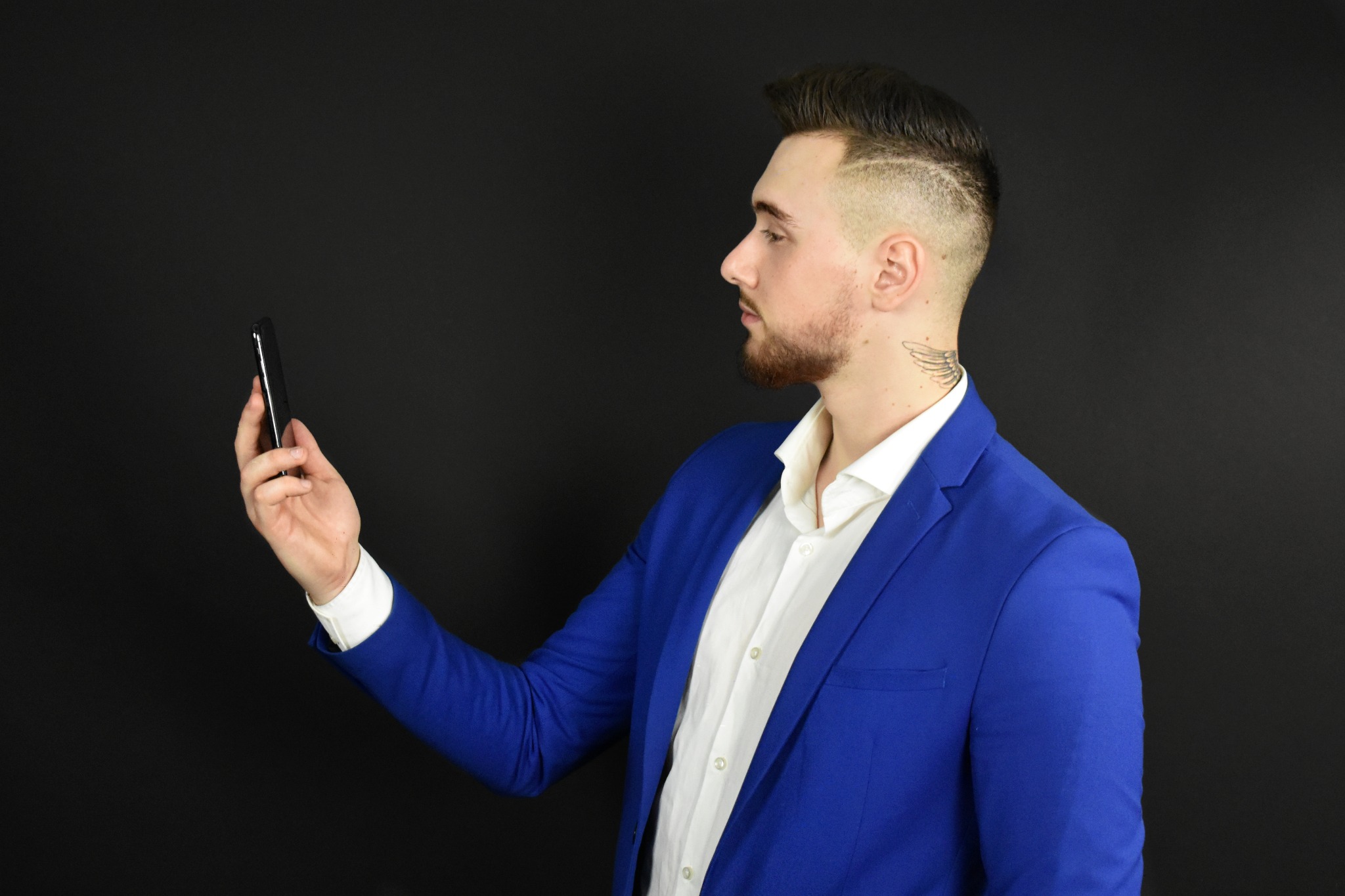 Mężczyzna w niebieskiej marynarce i białej koszuli, z tatuażem na szyi, trzyma telefon komórkowy na czarnym tle.