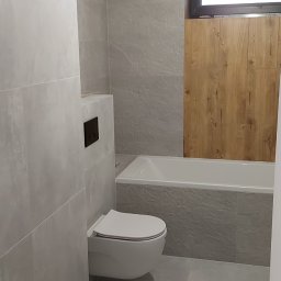 Fachbud - Wykończona łazienka z szarymi płytkami imitującymi kamień, podwieszana toaleta z czarnym przyciskiem, biała wanna obudowana płytkami, drewniana ściana obok okna, widoczne resztki po remoncie...