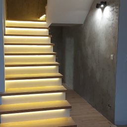 Fachbud - Nowoczesne schody z drewnianymi stopniami i subtelnym oświetleniem LED pod każdym stopniem, przylegające do ściany z szarą dekoracyjną strukturą, oświetlone dwoma reflektorami punktowymi.