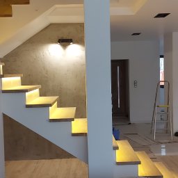 Fachbud - Nowoczesne wnętrze w trakcie wykańczania: podświetlane schody z drewnianymi stopniami, ściana z efektem betonu, surowe ściany w tle, widoczne narzędzia i materiały budowlane.