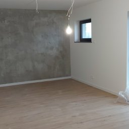 Fachbud - Surowe wnętrze po nałożeniu dekoracyjnej szarej gładzi na jedną ścianę, z widoczną podłogą z jasnego drewna, białymi ścianami i ościeżnicą okna, z niedokończonymi przewodami elektrycznymi...