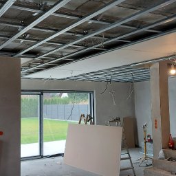 Fachbud - Wnętrze w trakcie wykończenia: metalowa konstrukcja sufitu podwieszanego, częściowo zamontowane płyty gipsowo-kartonowe, widoczne instalacje elektryczne, surowe ściany i schody, okna z widokiem...