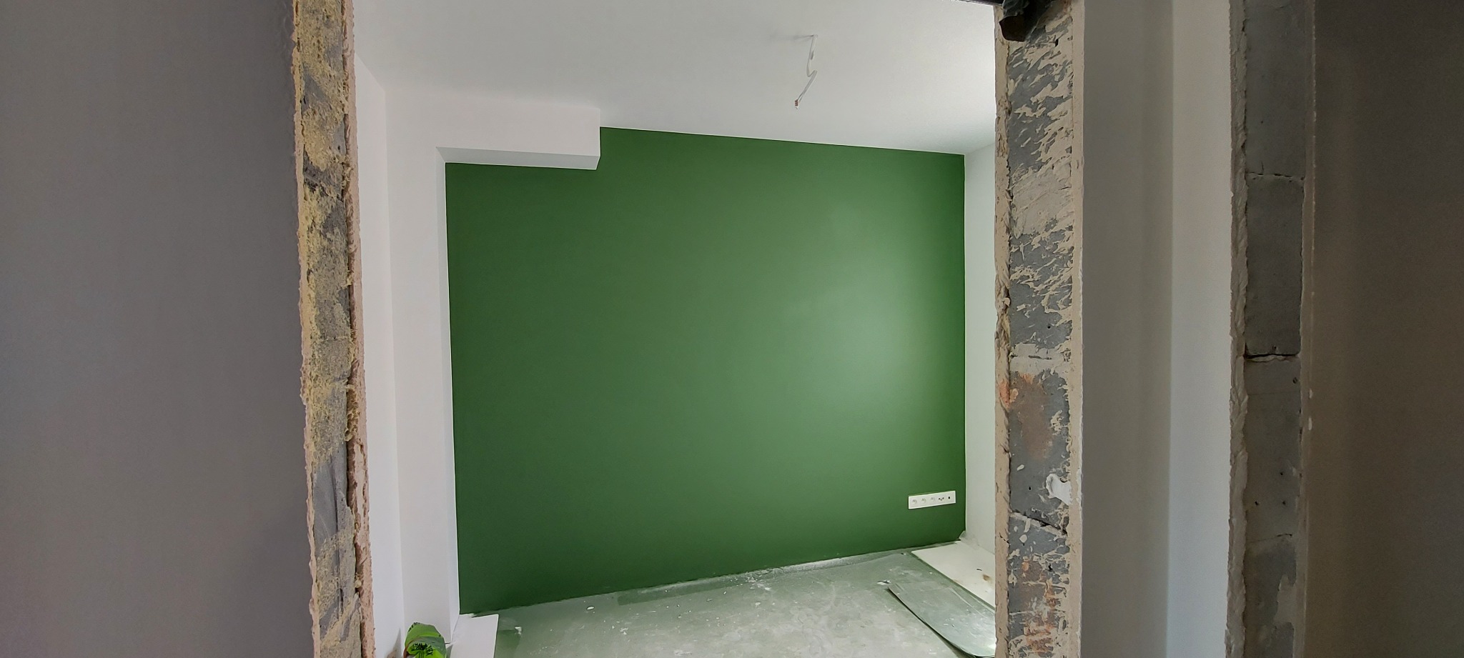 Świeżo pomalowane na oliwkowo-zielono ściany pokoju, widoczne przez surową ramę drzwi w trakcie remontu, z widocznymi przewodami elektrycznymi na suficie i gniazdkami na ścianie.