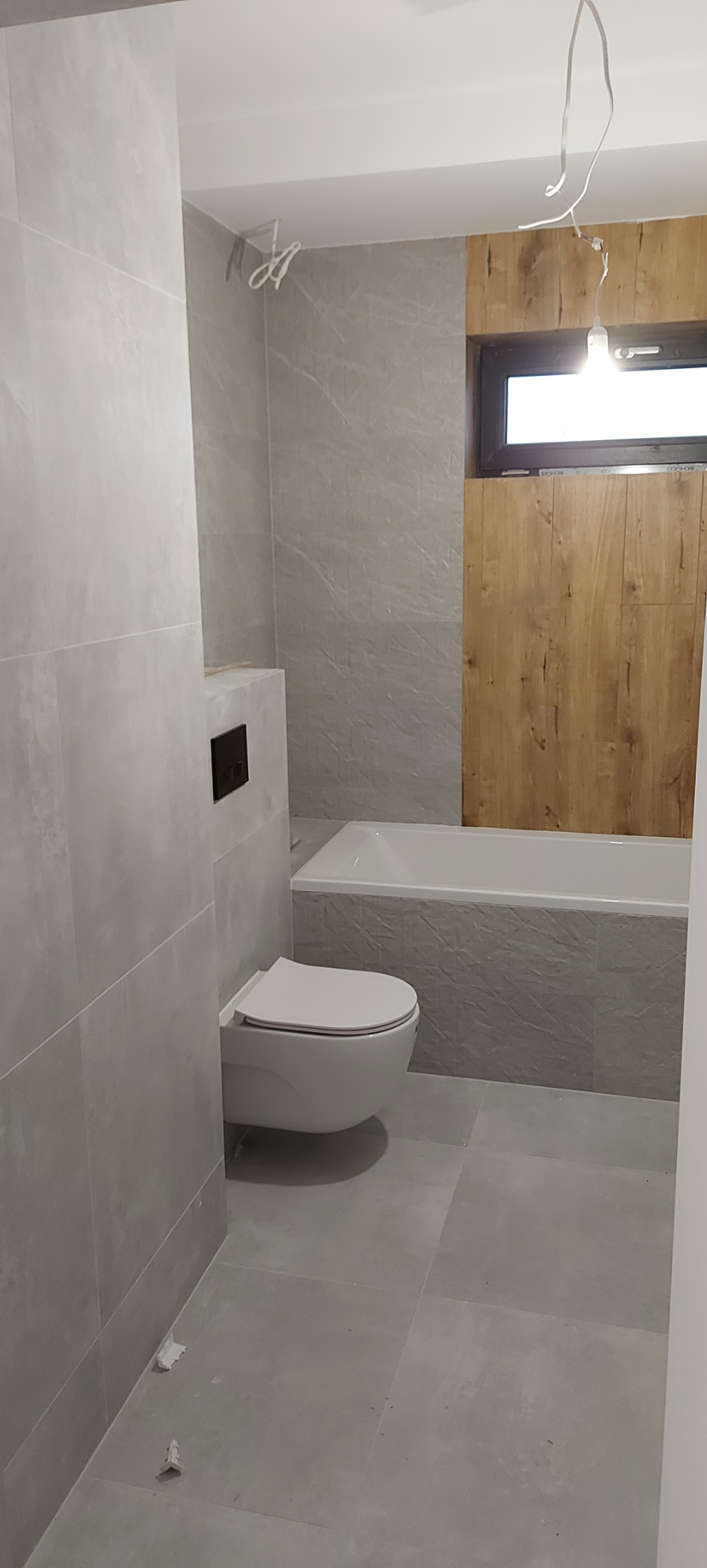 Wykończona łazienka z szarymi płytkami imitującymi kamień, podwieszana toaleta z czarnym przyciskiem, biała wanna obudowana płytkami, drewniana ściana obok okna, widoczne resztki po remoncie...