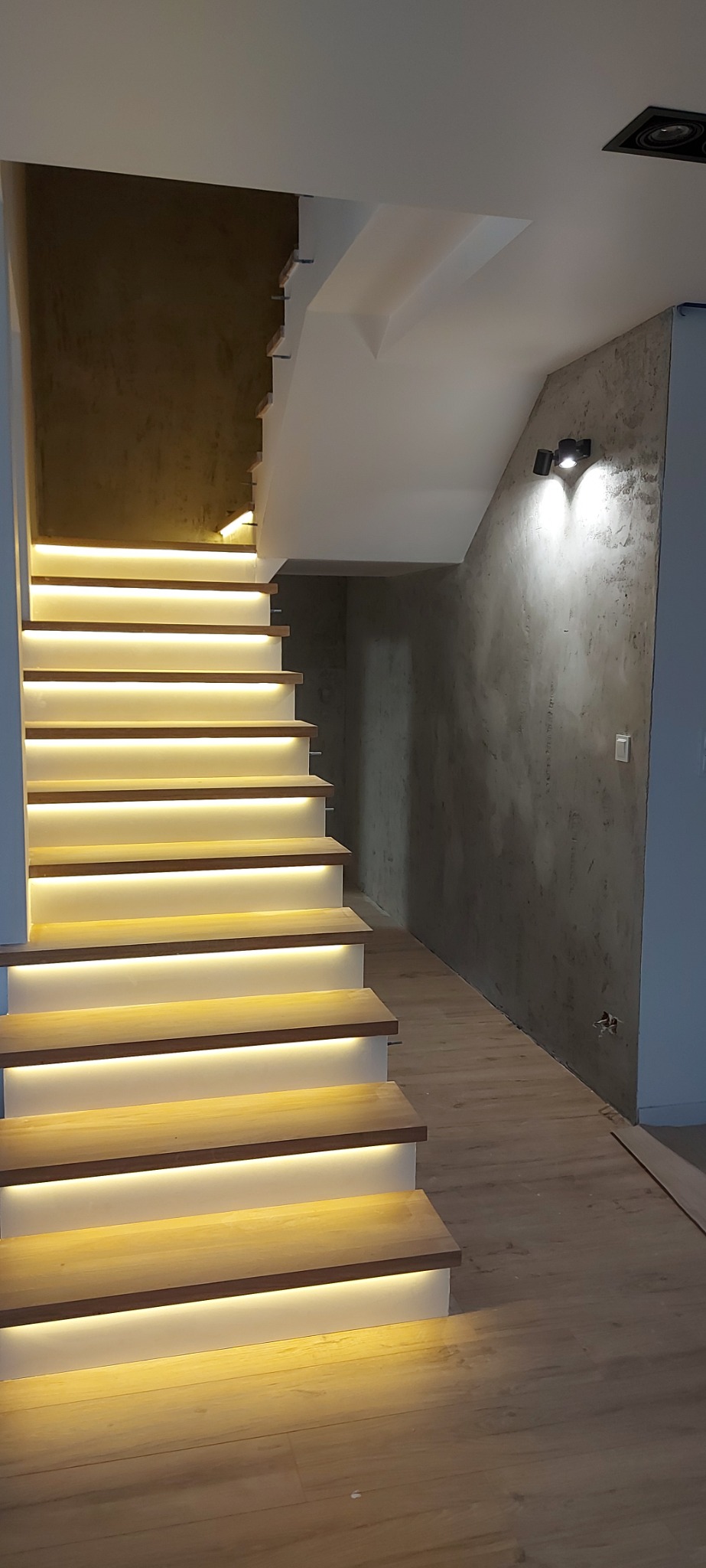 Nowoczesne schody z drewnianymi stopniami i subtelnym oświetleniem LED pod każdym stopniem, przylegające do ściany z szarą dekoracyjną strukturą, oświetlone dwoma reflektorami punktowymi.