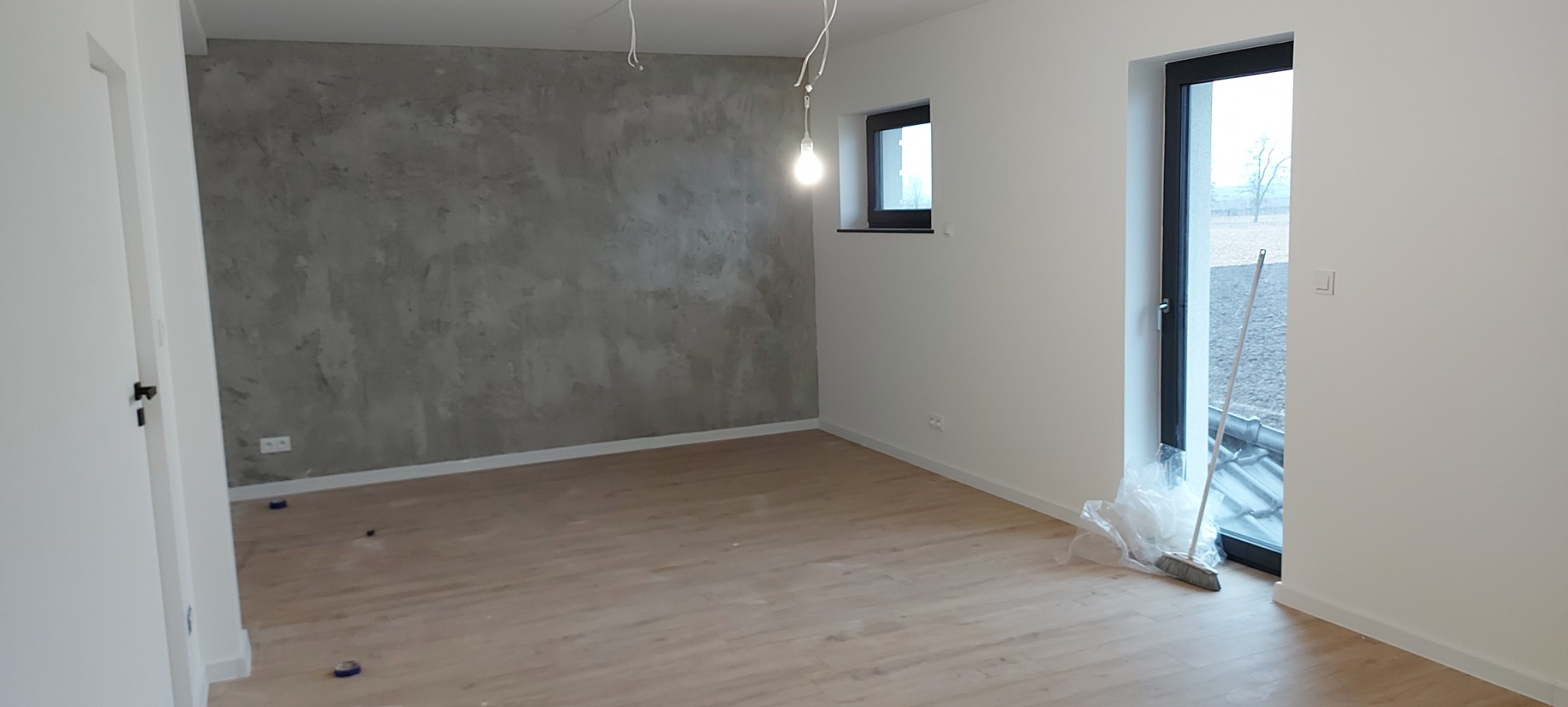 Surowe wnętrze po nałożeniu dekoracyjnej szarej gładzi na jedną ścianę, z widoczną podłogą z jasnego drewna, białymi ścianami i ościeżnicą okna, z niedokończonymi przewodami elektrycznymi...