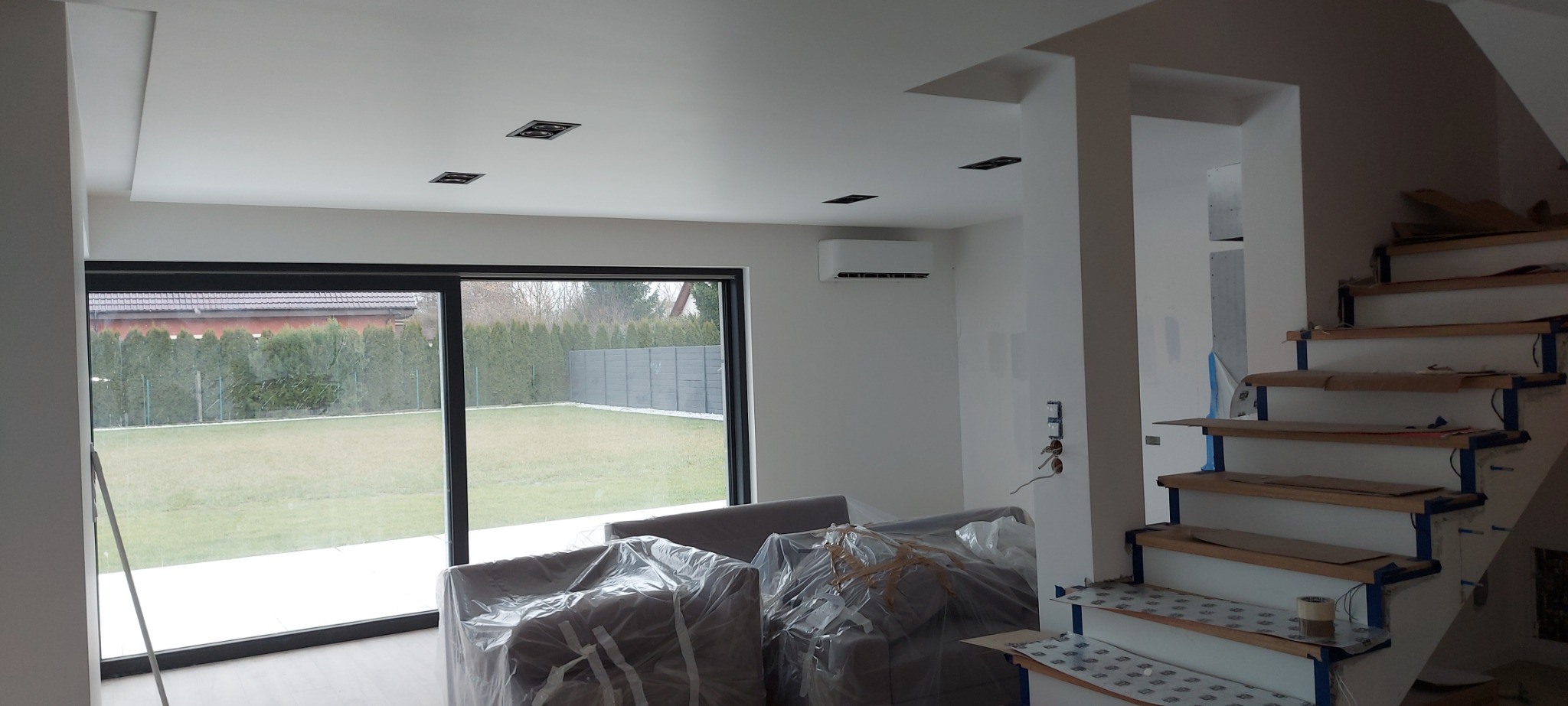 Wnętrze domu w trakcie prac wykończeniowych: świeżo pomalowane ściany na biało, zabezpieczone folią meble, schody z drewnianymi stopniami w trakcie montażu, widoczne puszki elektryczne i klimatyzator.