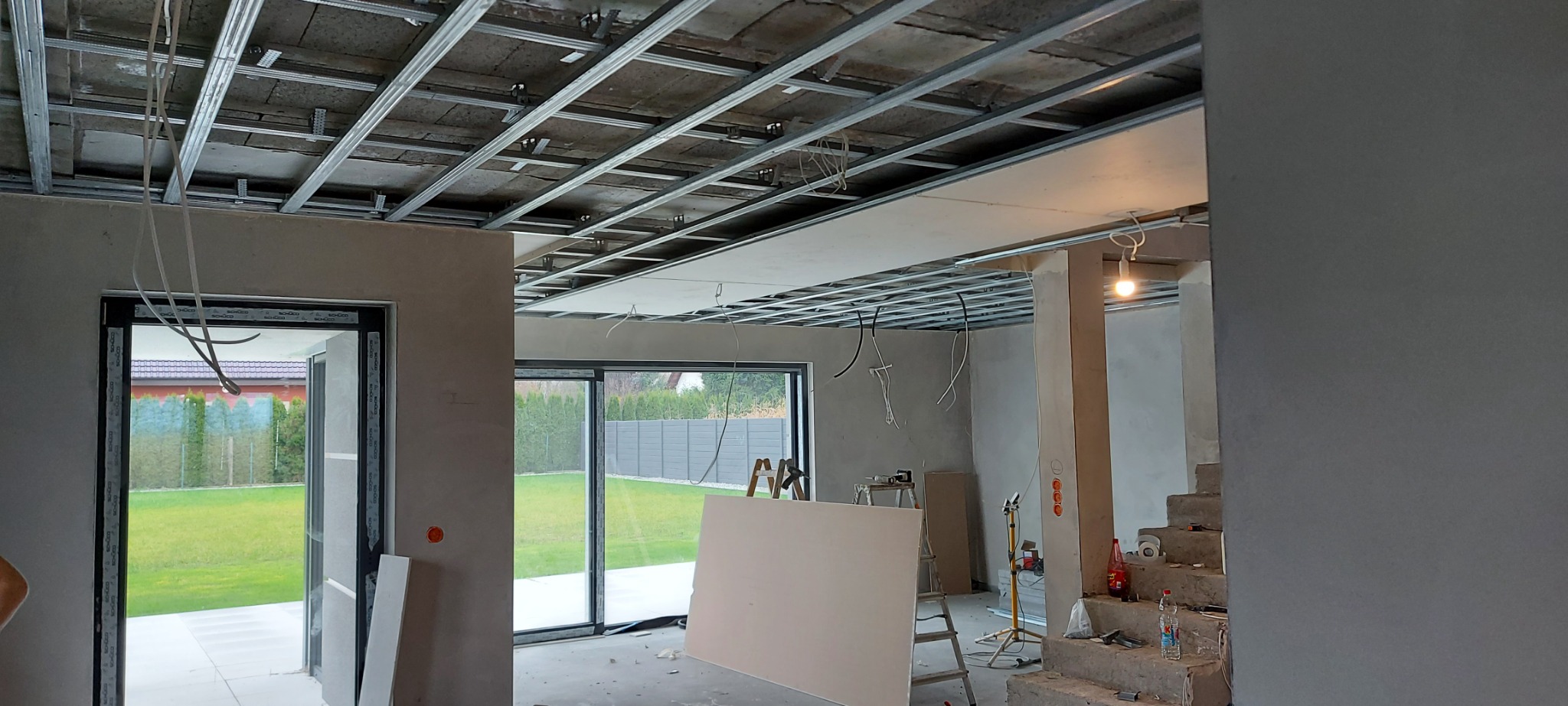 Wnętrze w trakcie wykończenia: metalowa konstrukcja sufitu podwieszanego, częściowo zamontowane płyty gipsowo-kartonowe, widoczne instalacje elektryczne, surowe ściany i schody, okna z widokiem...