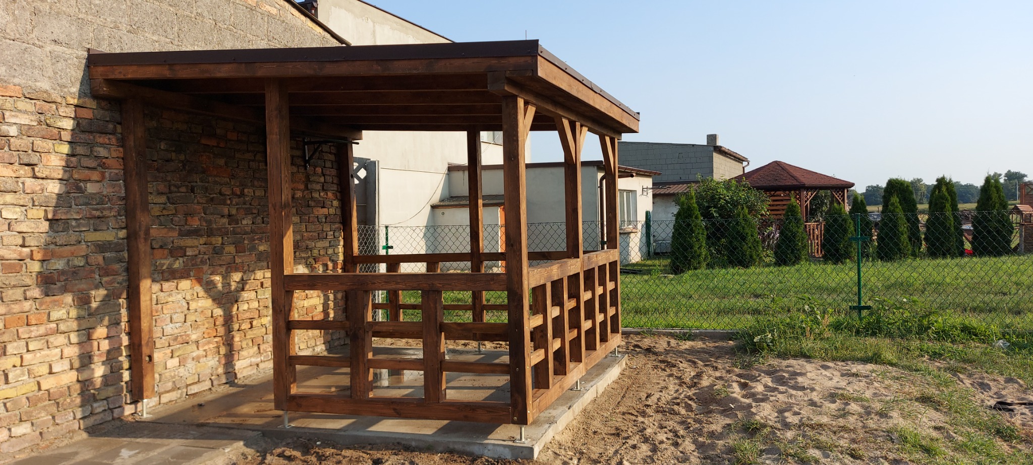 Drewniana altana z brązowym dachem, przylegająca do ceglanego budynku, widoczna z zewnątrz na tle trawnika i innych budynków.