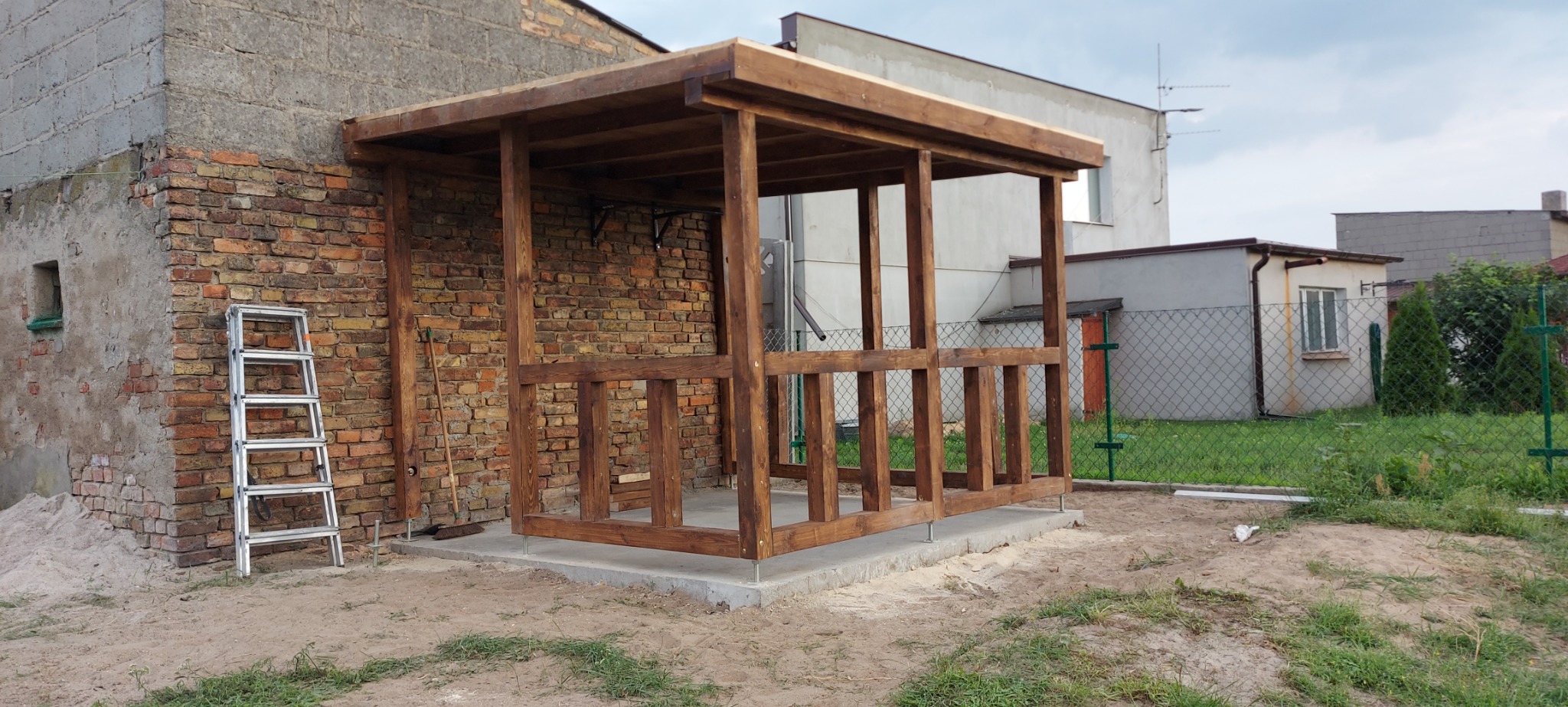 Drewniana konstrukcja zadaszenia tarasu przylegająca do ceglanego budynku, widoczna drabina aluminiowa i narzędzia ogrodnicze, teren wokół w trakcie prac ziemnych.