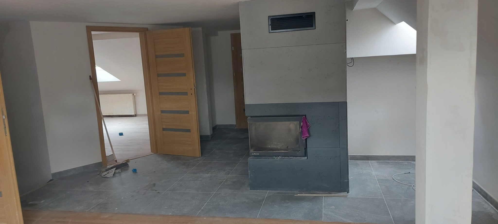 Wnętrze poddasza w trakcie remontu: szare płytki podłogowe, częściowo otwarte drewniane drzwi, kominek obłożony płytami z szarego betonu architektonicznego, surowe ściany przygotowane do dalszych...