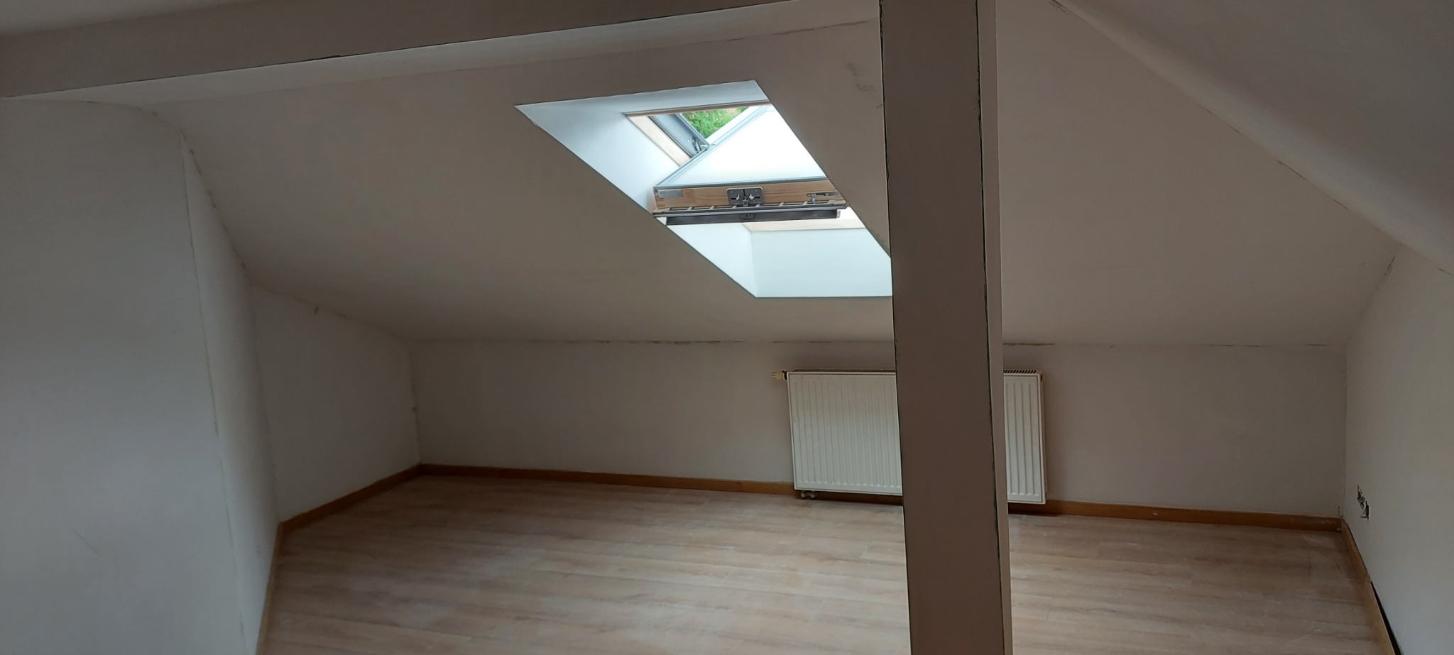 Poddasze po remoncie: jasne ściany, drewnopodobna podłoga, okno dachowe wpuszczające światło, widoczne grzejniki i pionowa belka konstrukcyjna.