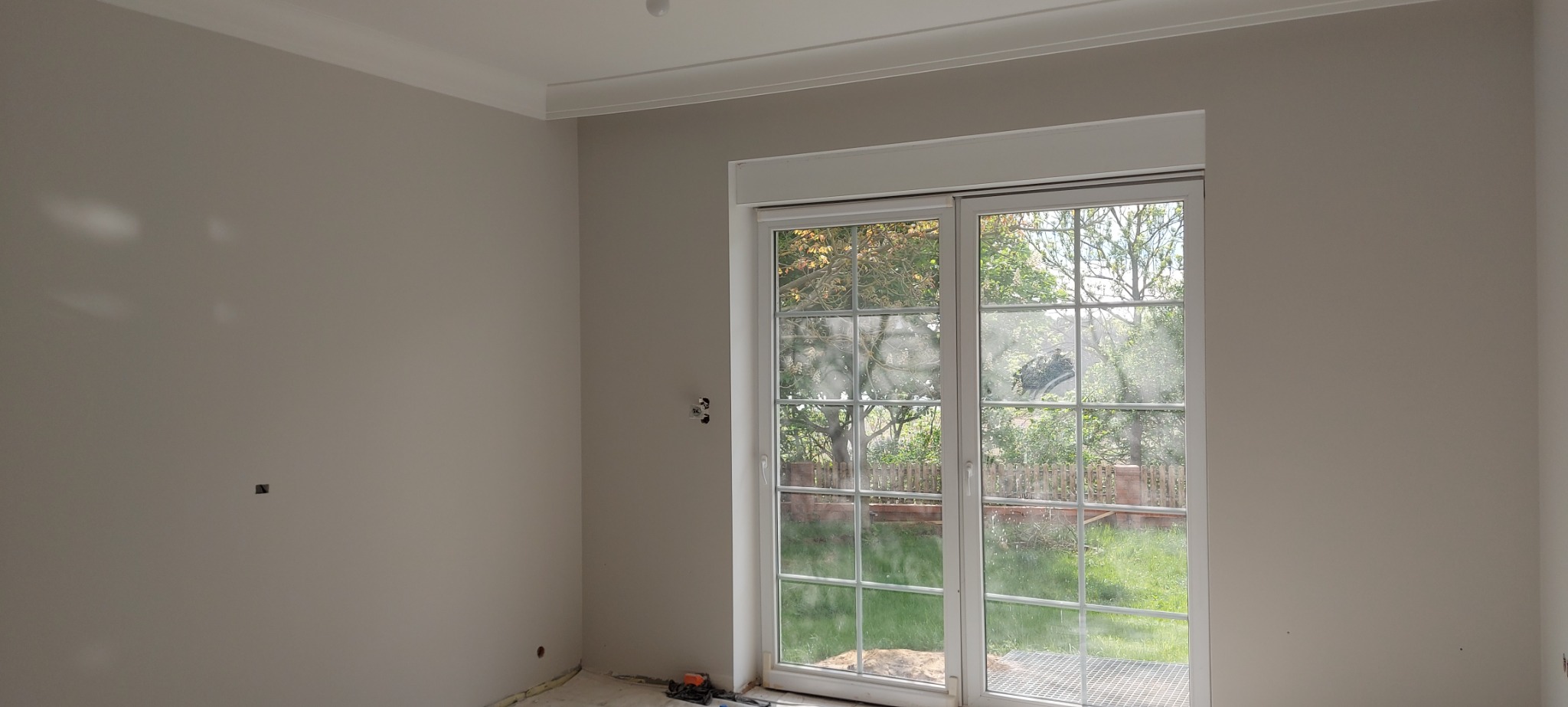 Wnętrze pokoju po remoncie, świeżo wykończone ściany w odcieniach szarości, białe listwy przysufitowe i duże okno przesuwne z widokiem na ogród, widoczne ślady prac na podłodze.