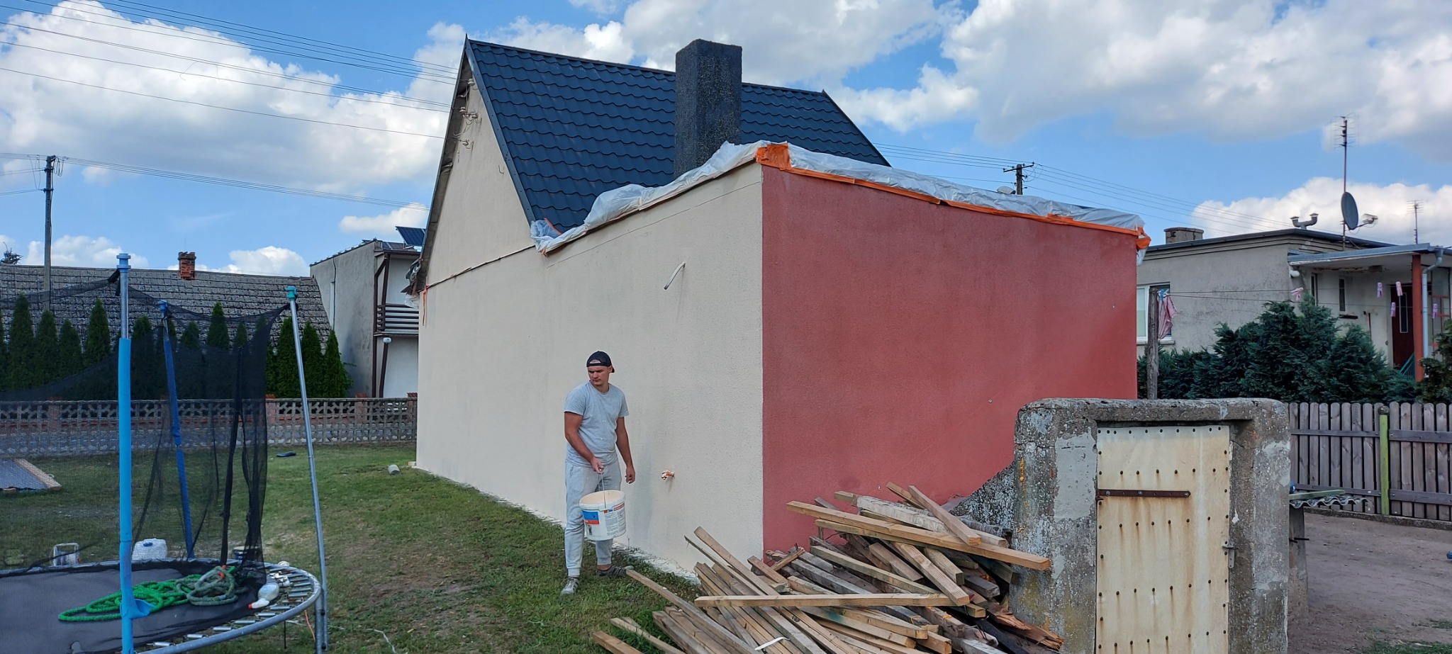 Mężczyzna z wałkiem i wiadrem farby maluje elewację budynku na dwa kolory, z widocznym przygotowaniem i zabezpieczeniem dachu folią.