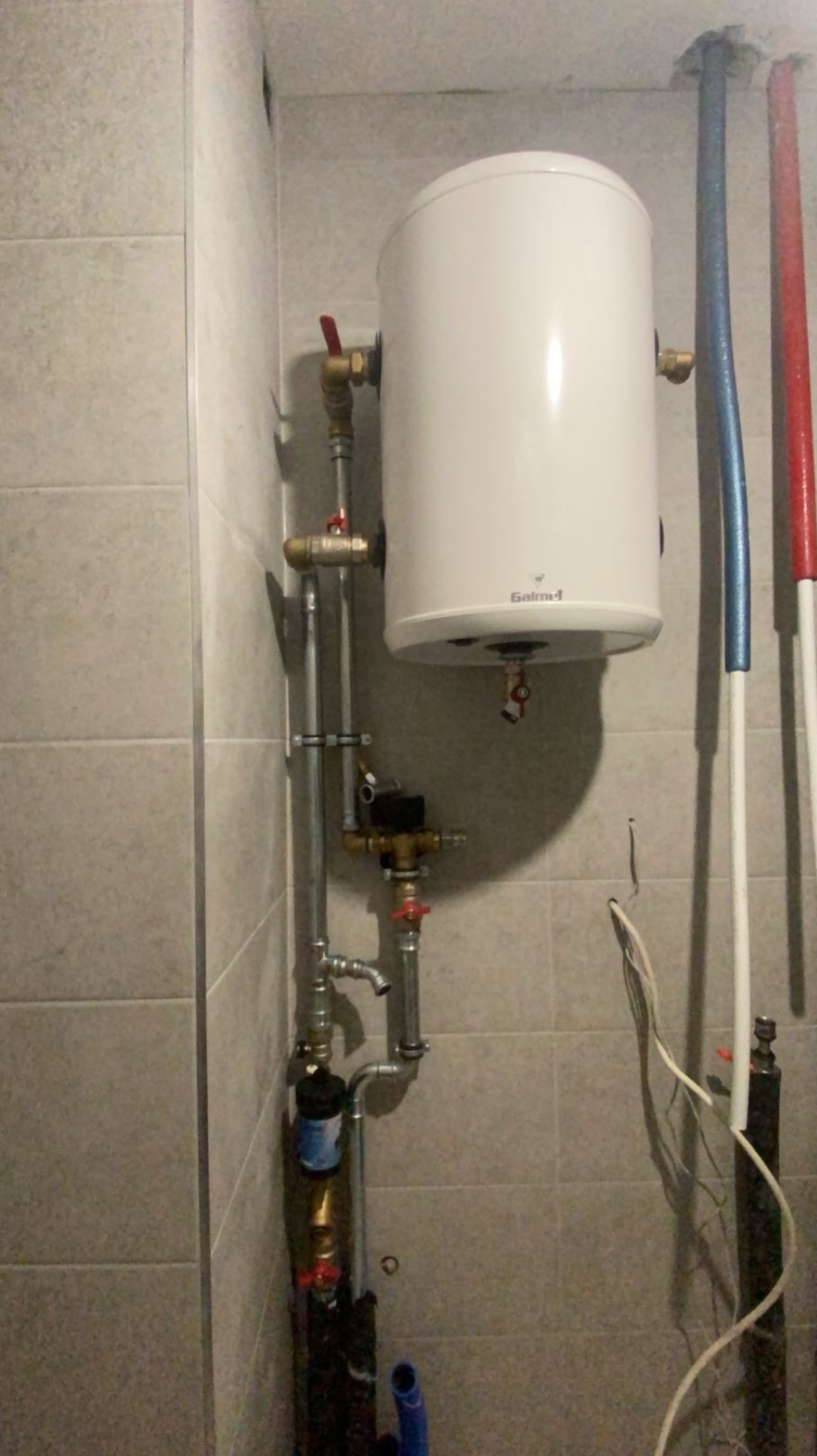 Bojler elektryczny Galmet wiszący na ścianie z widocznymi rurami doprowadzającymi wodę, zaworami i elementami instalacji hydraulicznej w pomieszczeniu wyłożonym płytkami.