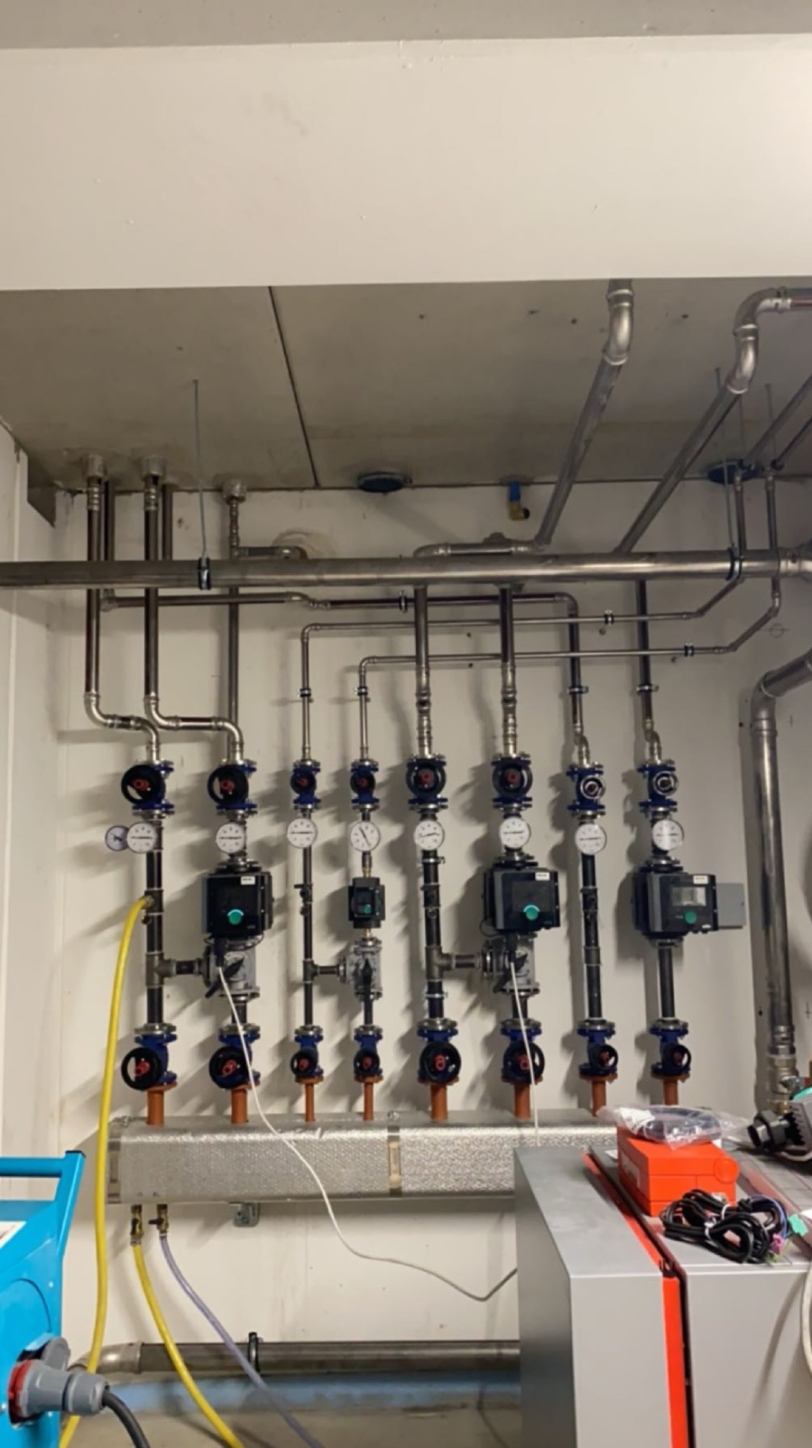 Rozdzielacz z licznymi zaworami i rurami w pomieszczeniu technicznym, widoczne manometry i niebieski wózek narzędziowy