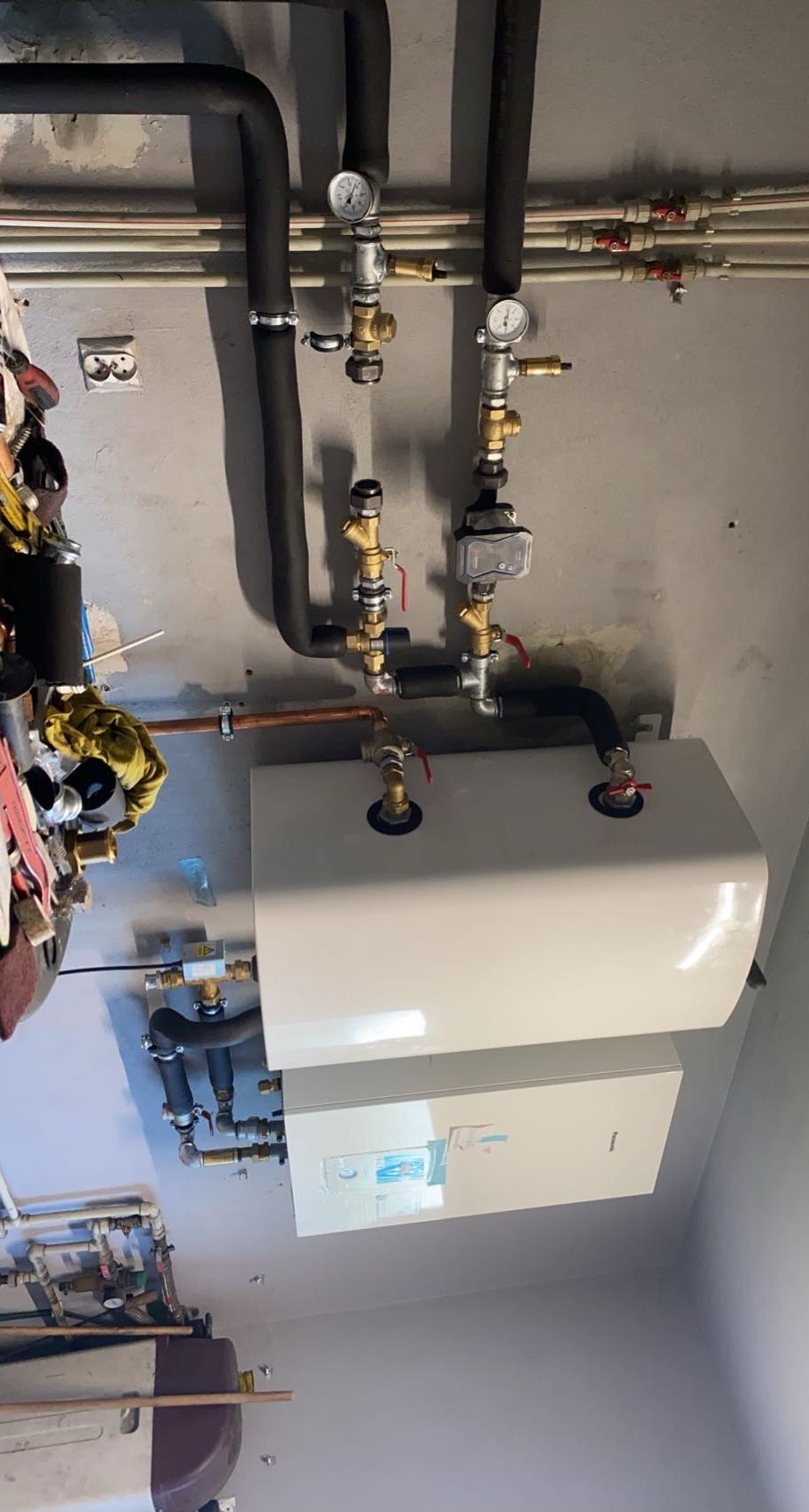 Nowoczesny piec grzewczy zamontowany na szarej ścianie, widoczne rury, zawory i manometry, instalacja w trakcie podłączenia, widoczny bałagan narzędzi.