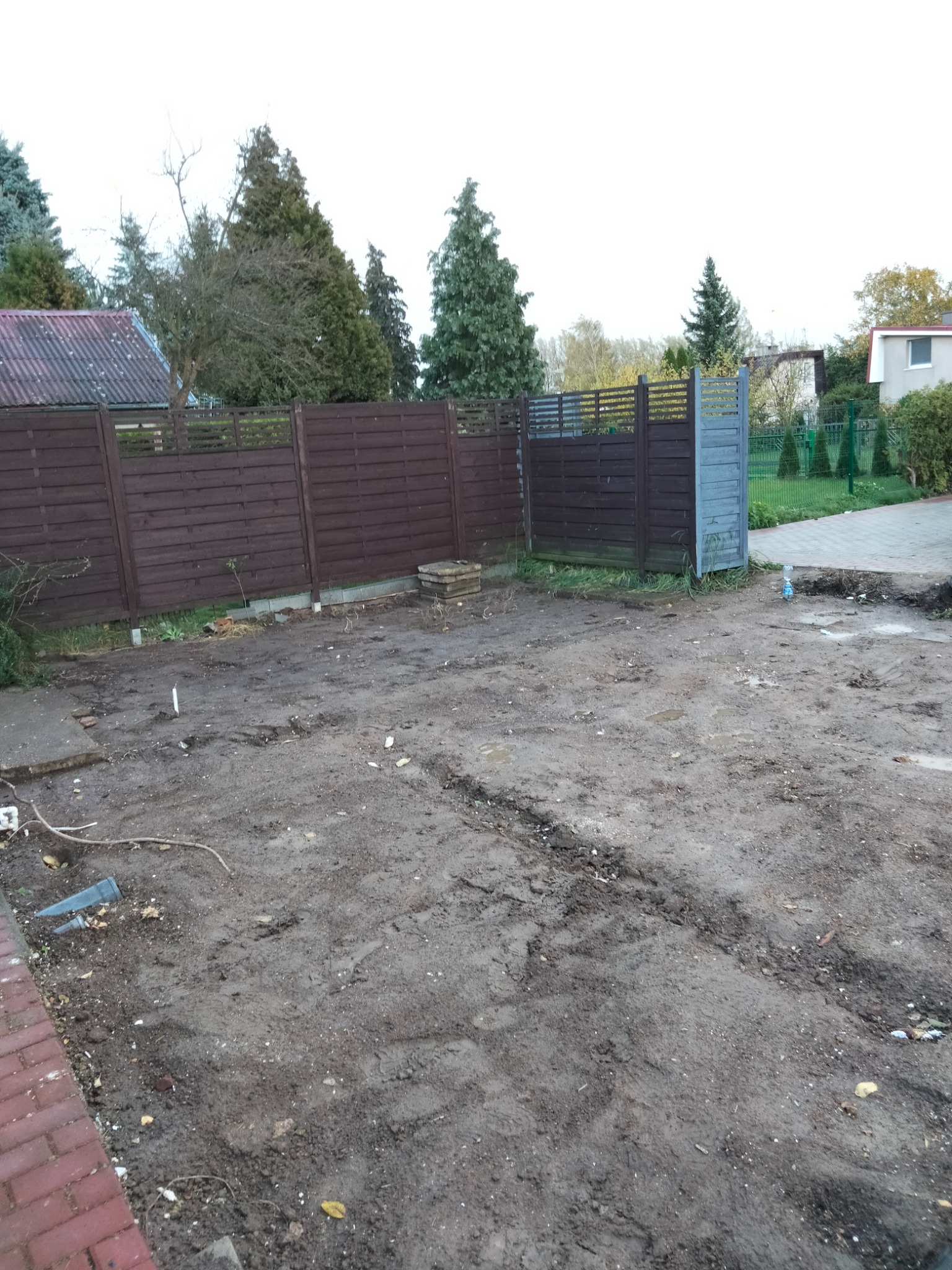 Teren po pracach ziemnych z widocznymi śladami opon, fragmentem ceglanego chodnika i drewnianym ogrodzeniem w tle.