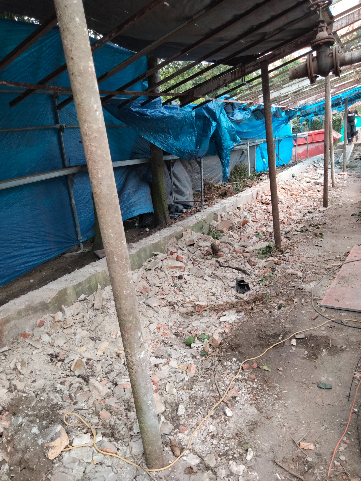 Fragment rozbiórki z widocznymi resztkami cegieł i gruzu na podłożu, zabezpieczonym rusztowaniem z niebieską plandeką w tle.