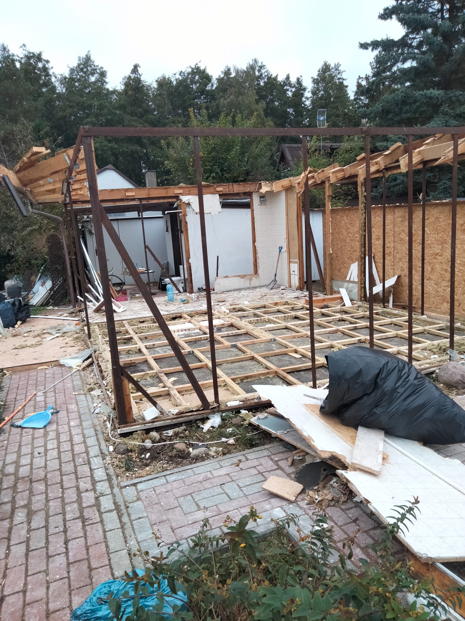 Częściowo rozebrany budynek z metalową konstrukcją szkieletową, widoczne drewniane elementy podłogi, gruz i śmieci na zewnątrz, w tle drzewa.