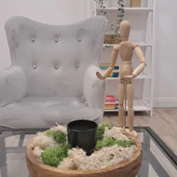 Jasnoszary fotel uszak, drewniany manekin na szklanym stoliku z dekoracją z mchu i czarnej doniczki, w tle biały regał z rośliną pnącą i dekoracyjną głową.