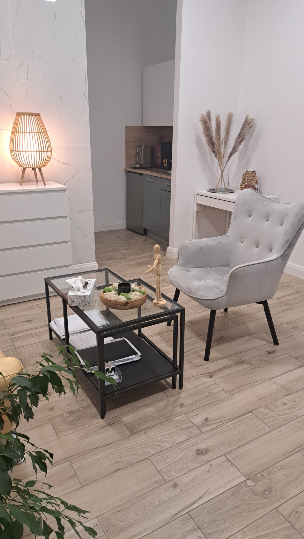 Jasne wnętrze gabinetu z szarym fotelem, stolikiem kawowym z drewnianą dekoracją, lampą z wikliny i elementami wystroju w stylu skandynawskim.