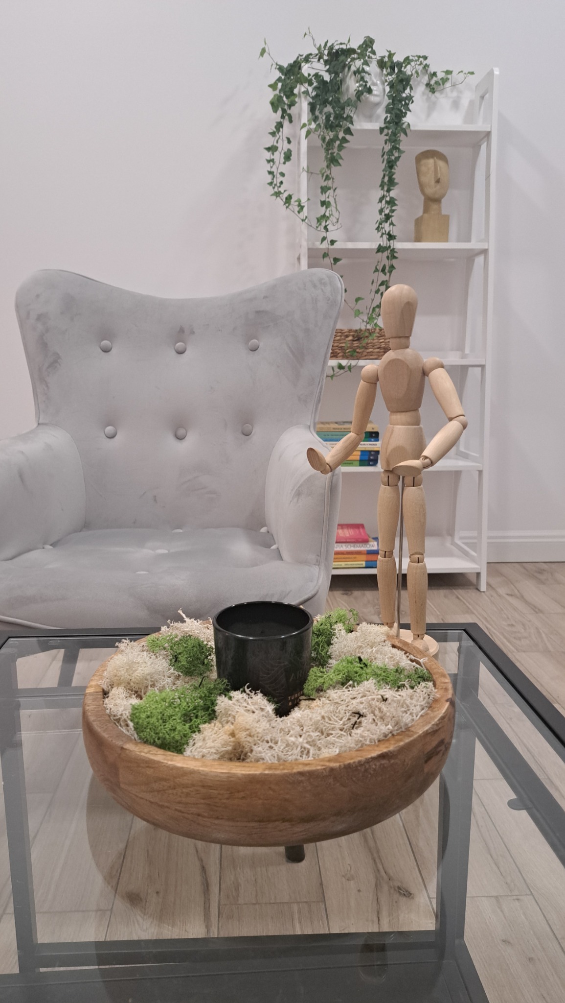 Jasnoszary fotel uszak, drewniany manekin na szklanym stoliku z dekoracją z mchu i czarnej doniczki, w tle biały regał z rośliną pnącą i dekoracyjną głową.