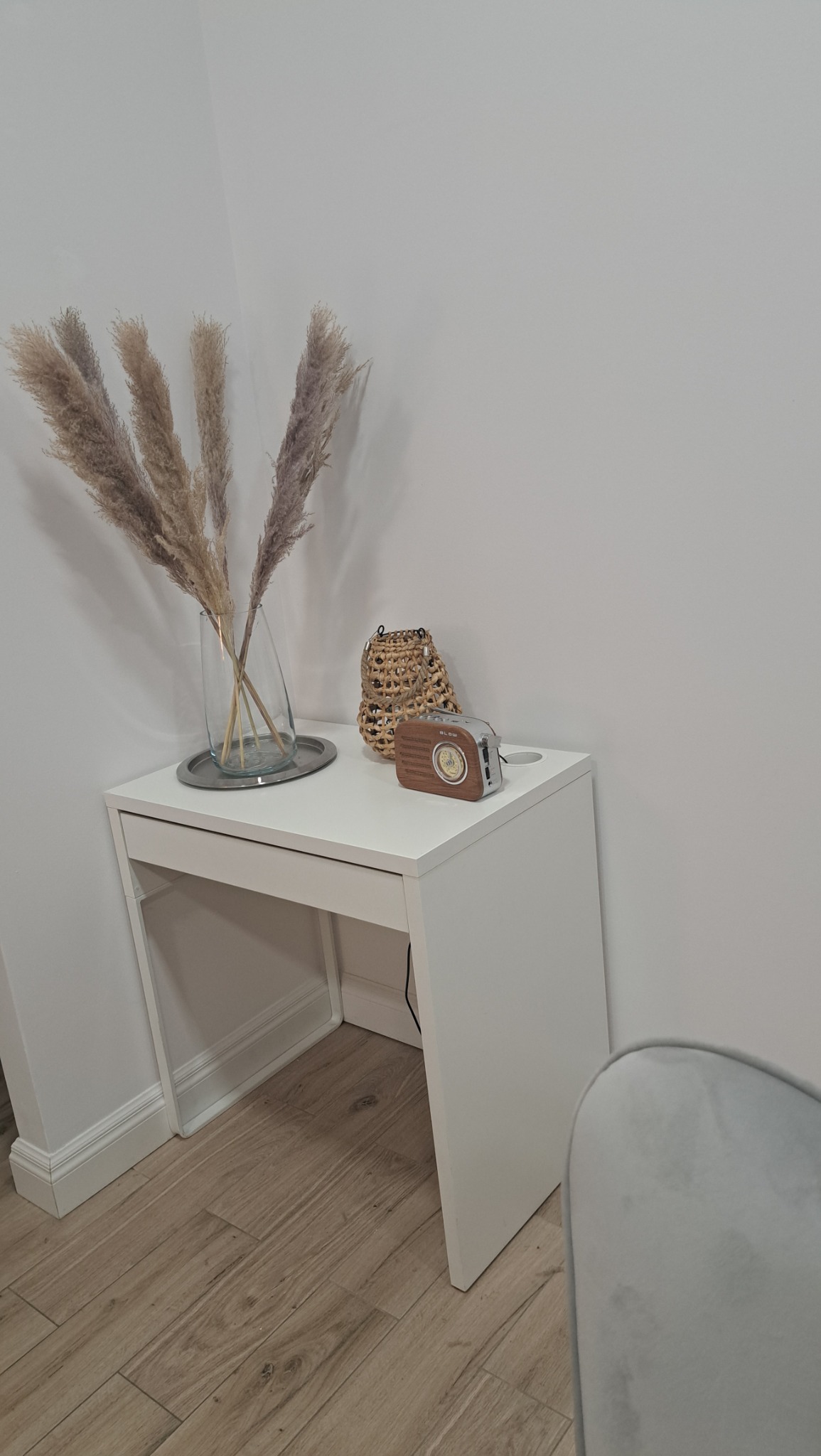 Minimalistyczny kącik: białe biurko z pampasami w szklanym wazonie na srebrnej tacy, wiklinowy koszyk i retro radio Blow, ustawione przy białej ścianie i drewnianej podłodze.