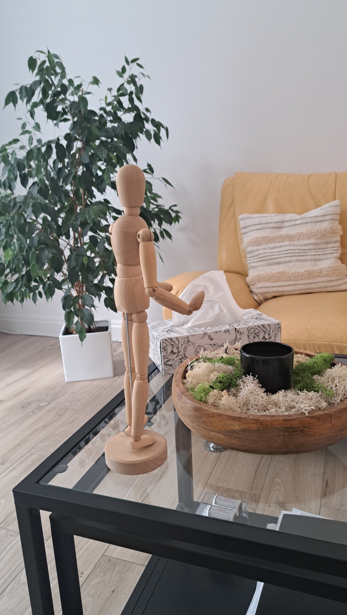 Drewniany model człowieka wyciąga rękę w stronę pudełka chusteczek na szklanym stoliku kawowym obok dekoracyjnej misy z mchem, z żółtą kanapą i rośliną w tle.