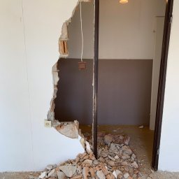 Atlantis b&b - Uszkodzona ściana z widocznymi cegłami i instalacją elektryczną obok futryny drzwiowej, gruz na podłodze, widok na sąsiednie pomieszczenie z lampą sufitową.