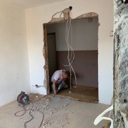 Atlantis b&b - Pracownik w trakcie wykuwania otworu drzwiowego w ścianie podczas prac remontowych, widoczne gruz i narzędzia.