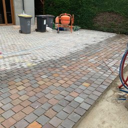 Atlantis b&b - Układanie kostki brukowej zróżnicowanej kolorystycznie na podjeździe, widoczna betoniarka, ślady po wytyczaniu terenu, w tle dom jednorodzinny.