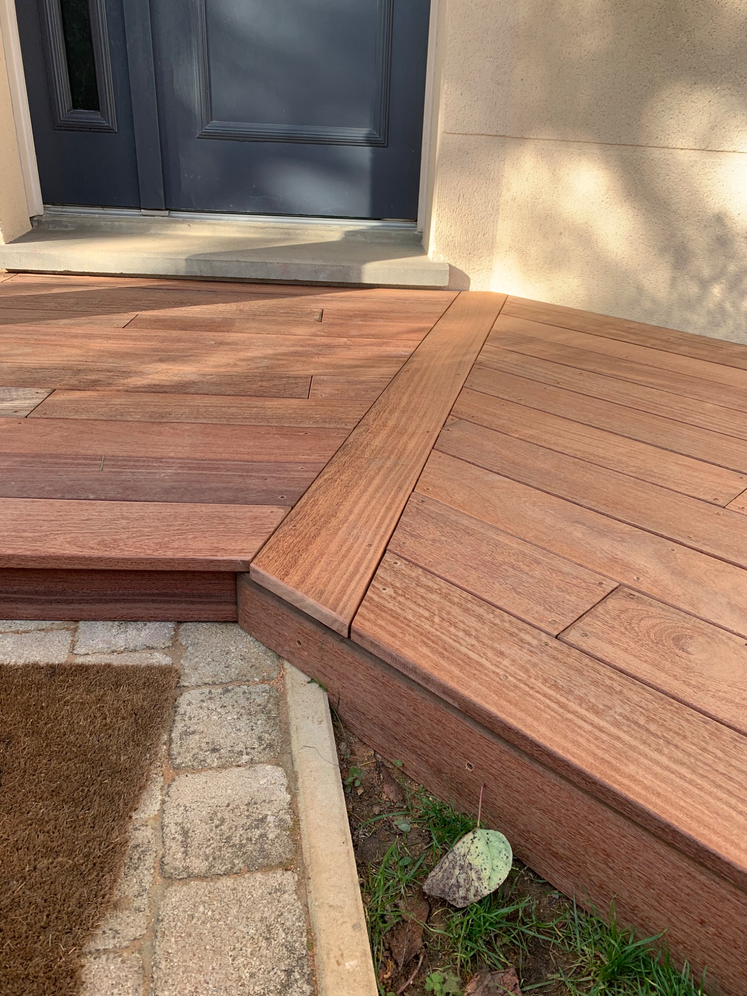 Nowoczesny podest wejściowy z kompozytowych desek w kolorze ciepłego brązu, prowadzący do ciemnych drzwi wejściowych, z widocznym fragmentem wycieraczki i kostki brukowej.