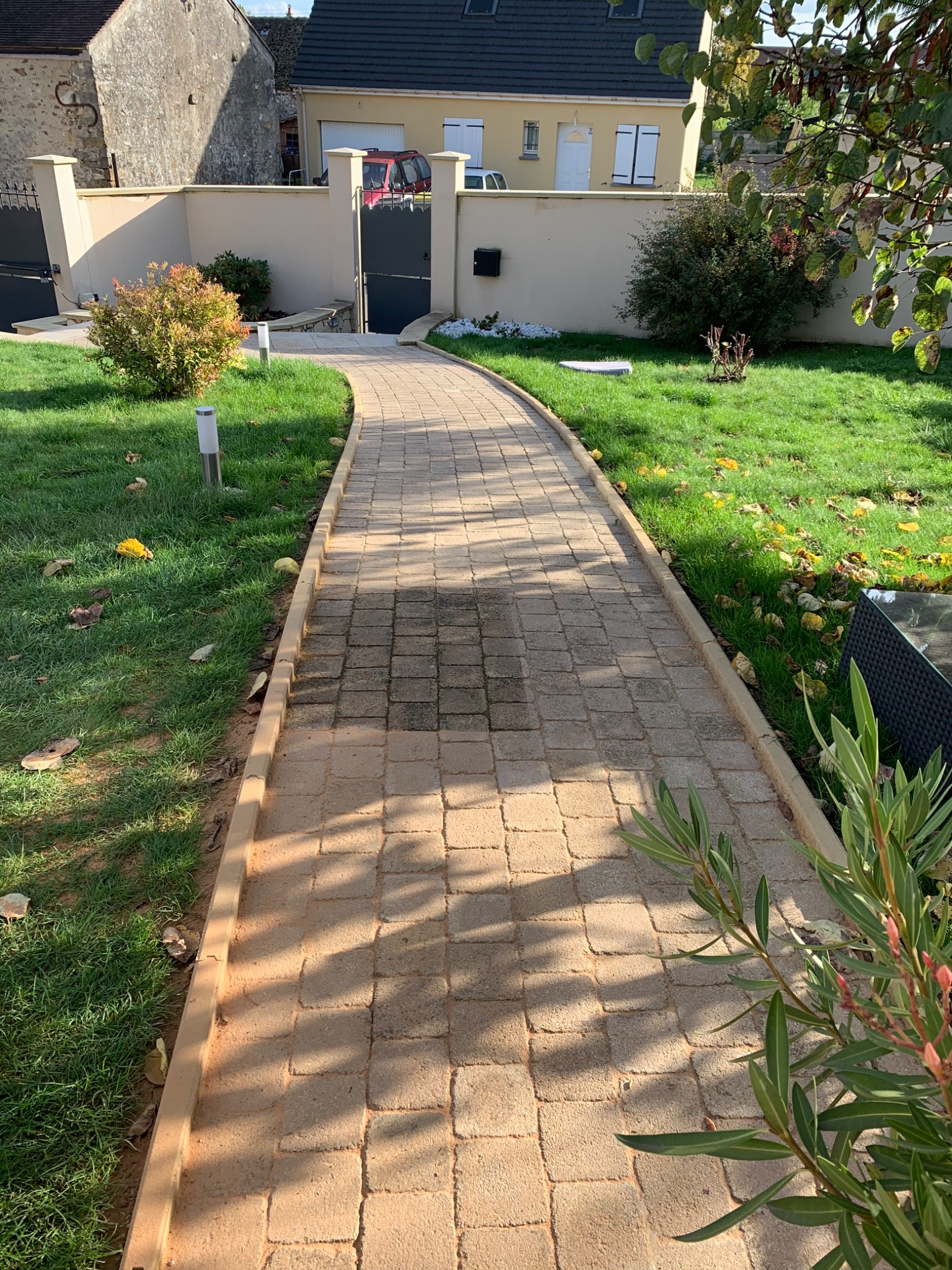Kwadratowa ścieżka z kostki brukowej prowadząca przez trawnik w kierunku bramy posesji, z widocznym fragmentem domu w tle.