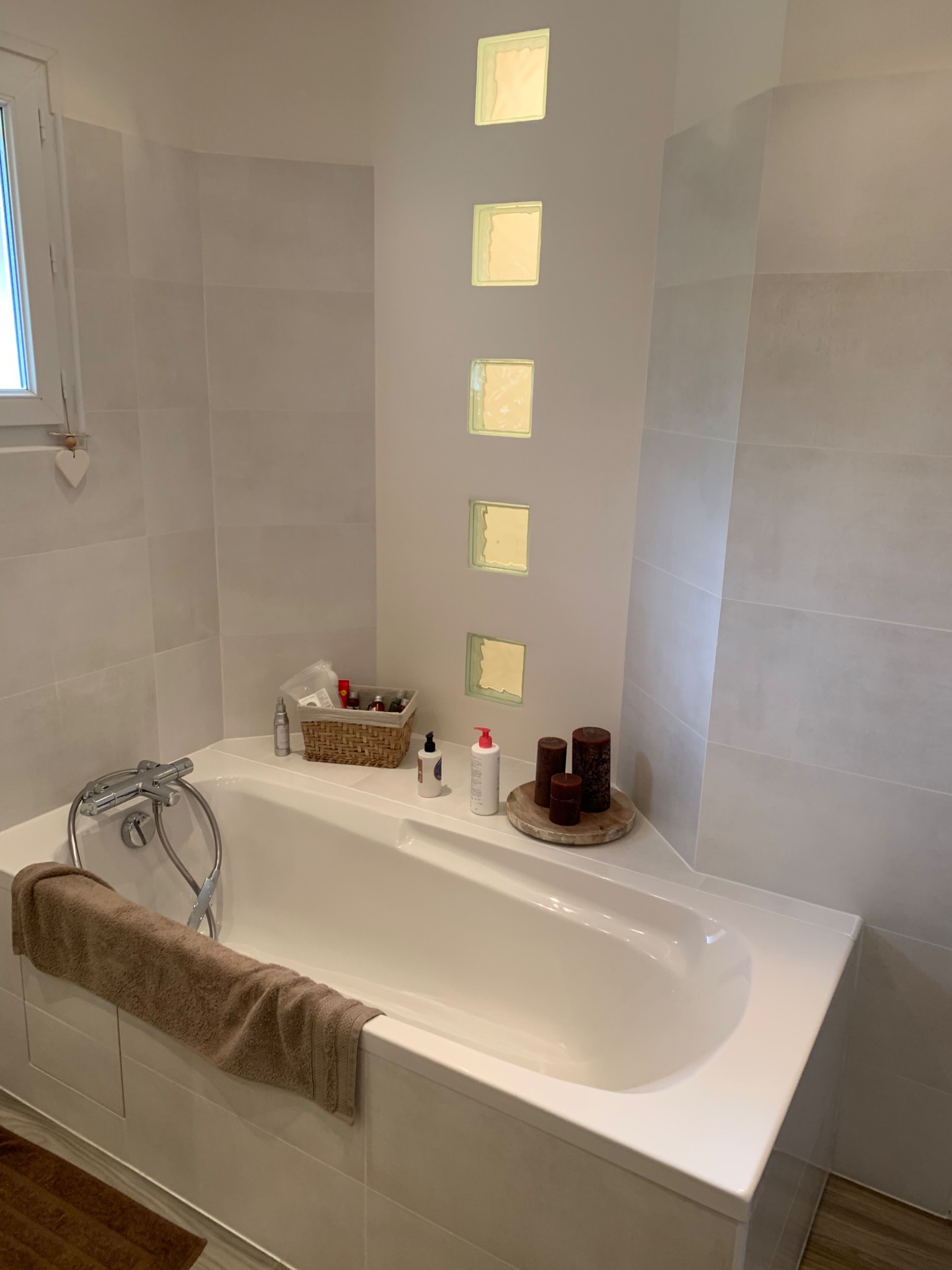 Jasna łazienka z wanną obudowaną płytkami, szklane luksfery w ścianie, wiklinowy koszyk z kosmetykami i brązowy ręcznik na wannie.